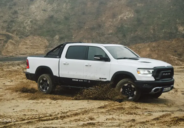 Biały Dodge Ram 1500 z czarnymi dodatkami pokonuje piaszczysty teren, wzbijając błoto.