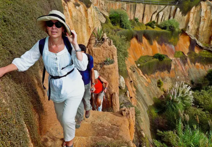 Hikers ascend a rocky path during hiking trips in Portugal, with lush green vegetation clinging to the orange cliffs.