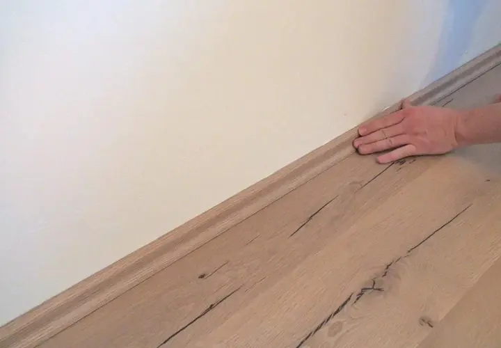 Hand drückt Sockelleisten an die Wand. Der Boden ist aus hellem Holz.