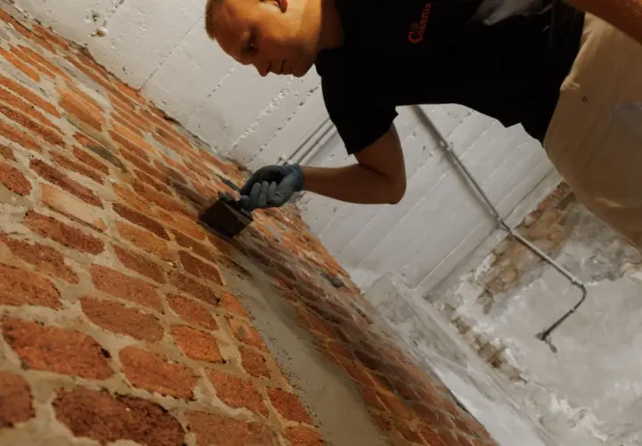Mann mit Handschuhen trägt braune Wanne im Keller auf Ziegelwand auf.