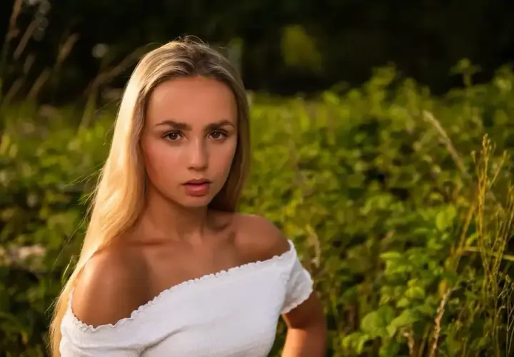 Piękna fotografia portretowa młodej kobiety z długimi blond włosami, w białej bluzce, na tle zielonej natury.