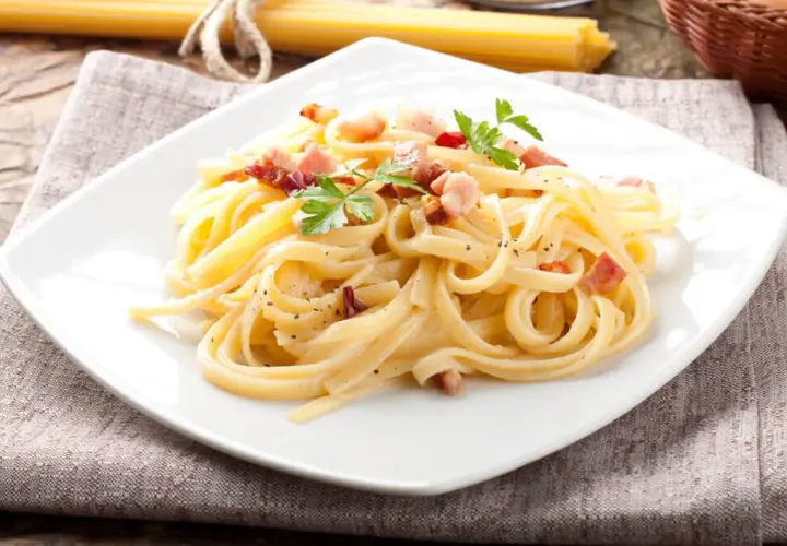Pyszne spaghetti carbonara z boczkiem i natką pietruszki na białym talerzu, obok surowy makaron.