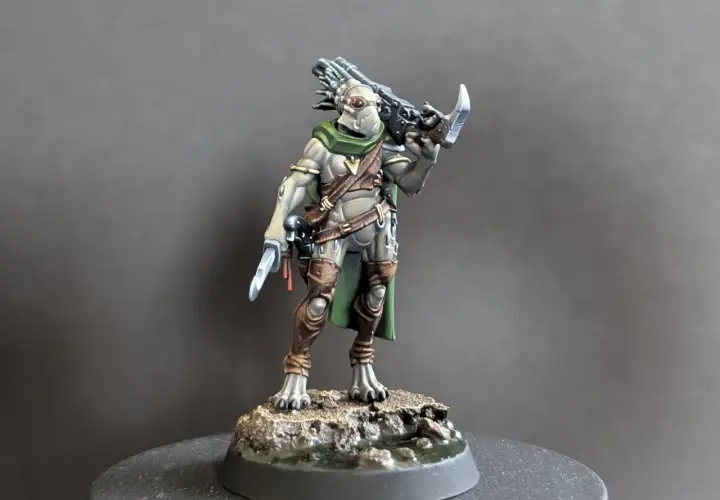 Zbrojny kroot w zielonej pelerynie i brązowych ochraniaczach, z nożem w dłoni i karabinem na plecach, gotowy do walki.