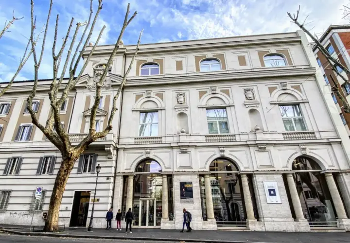 Le **Palazzo Merulana**, un édifice élégant, se dresse sous un ciel bleu nuageux, orné d'arbres dénudés aux branches graphiques.
