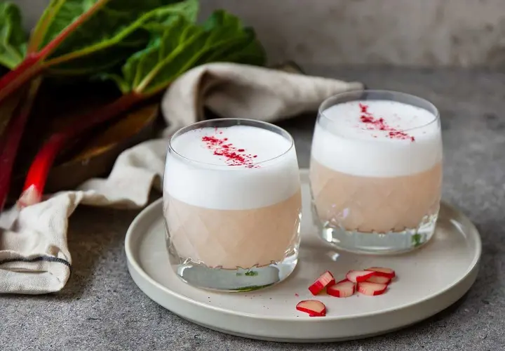 Två glas med en krämig rabarber sour, toppad med skum och strödd med torkad rabarber.