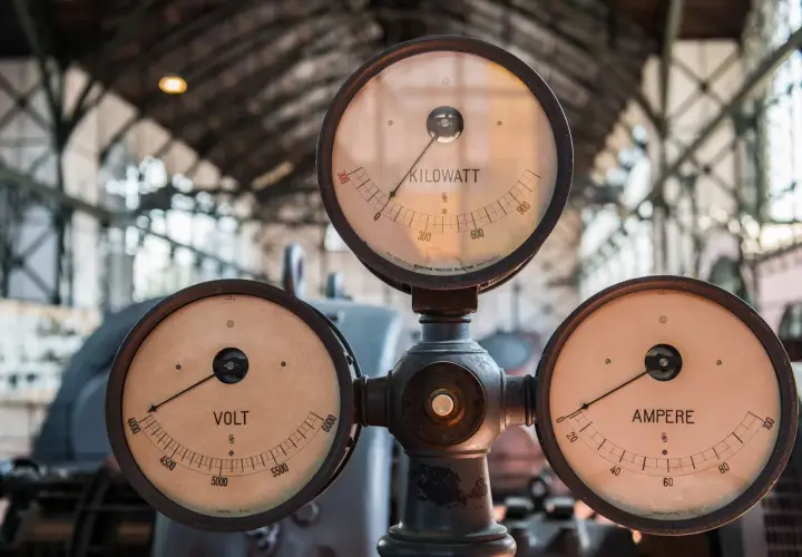 Trzy zabytkowe mierniki: VOLT, AMPERE i KILOWATT, wskazujące wartości prądu i mocy.