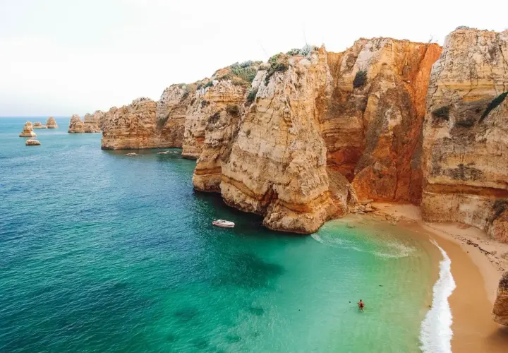 Aguas turquesas y acantilados dorados en las **playas Algarve**. Una pequeña barca navega cerca de una cala escondida donde una persona nada.