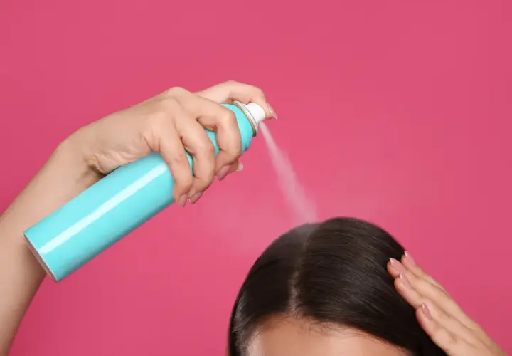 Dłoń trzymająca turkusowy spray z suchym szamponem, rozpylająca go na ciemne włosy.
