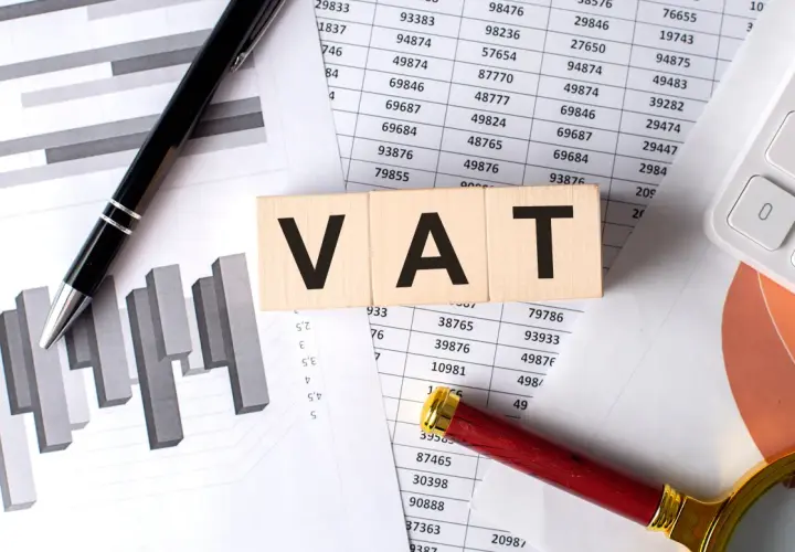Drewniane klocki z napisem VAT leżą na dokumentach z wykresami i tabelami. Jak odliczyć VAT z faktury? Pióro i lupa sugerują analizę.