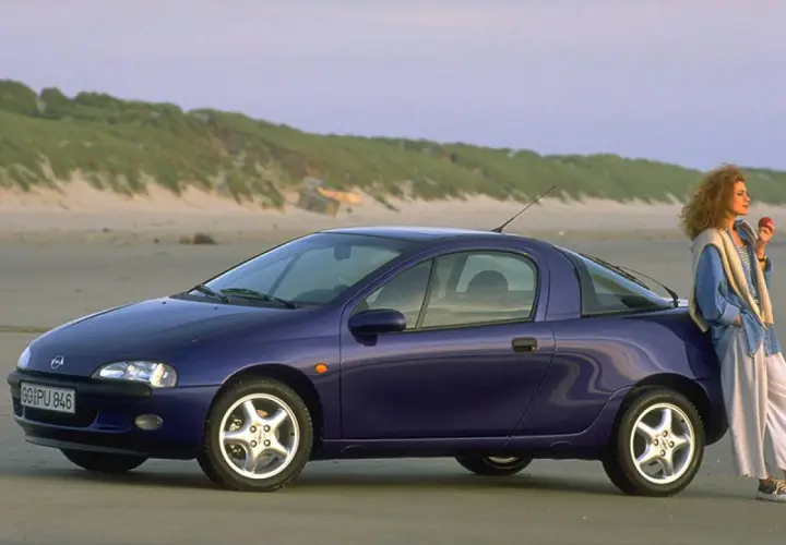 Una donna appoggiata a una Opel Tigra blu, icona delle macchine anni 90, sulla spiaggia al tramonto.