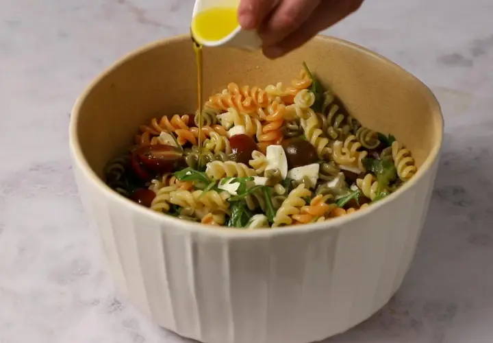 Un bol lleno de pasta de colores, tomates cherry, queso y rúcula, con aceite de oliva vertiéndose sobre ella.