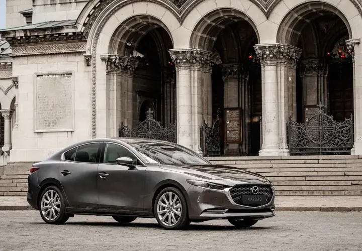 Elegancka, szara Mazda 3 2025 przed monumentalną, kamienną budowlą z arkadami i ozdobnymi bramami.
