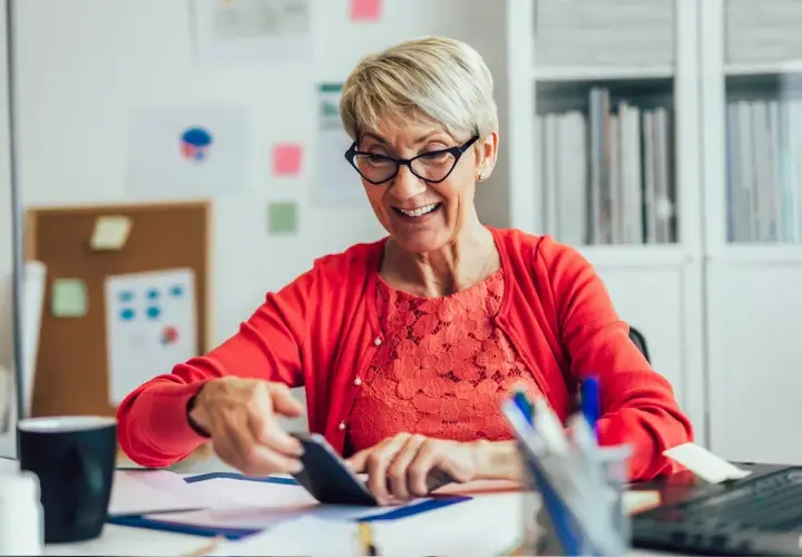 Uśmiechnięta starsza pani przy biurku sprawdza coś na telefonie, zastanawiając się, ile można dorobić do emerytury.
