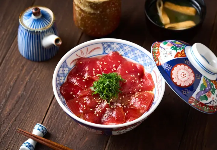 A bowl of delicious Japanese sweet rice topped with thinly sliced tuna, sesame seeds, and green garnish. A decorative lid rests beside it.