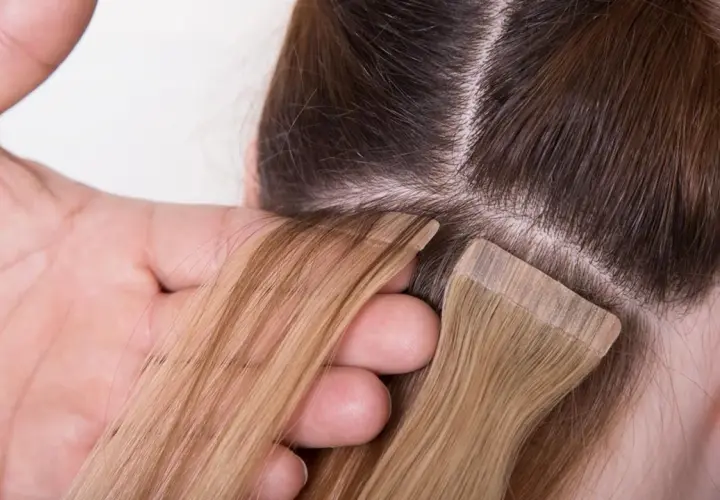Manos aplicando extensiones adhesivas rubias a cabello castaño. El proceso de colocación de extensiones adhesivas es delicado.