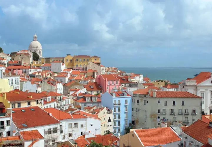 One day in Lisbon, a panorama of colorful buildings with red-tiled roofs stretches towards the shimmering blue sea under a cloudy sky.