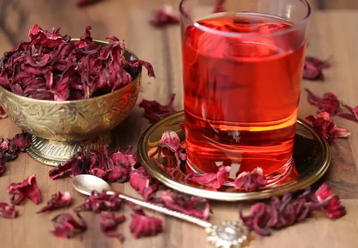 Ein Glas leuchtend roter Malva Sylvestris Tee steht neben einer Schale getrockneter Blüten. Getrocknete Blütenblätter sind auf dem Holztisch verstreut.