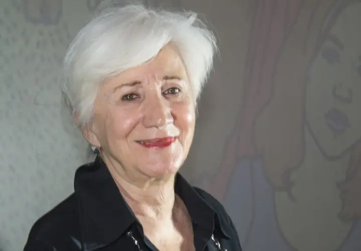 Olympia Dukakis ler vänligt mot kameran, med sitt karakteristiska vita hår och en svart skjorta. Bakom henne anas en tecknad figur.