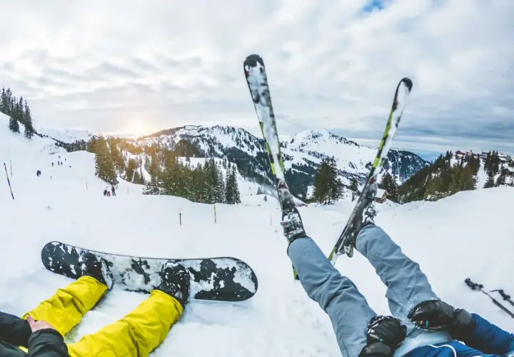 Dwie osoby odpoczywają na śniegu. Jedna leży na desce snowboardowej, druga ma założone narty. W tle góry i stoki narciarskie.