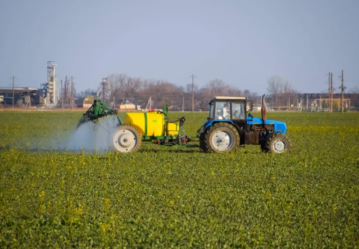 Niebieski traktor z żółtym zbiornikiem wykonuje oprysk na polu zielonych roślin. W tle widać przemysłowe budynki.