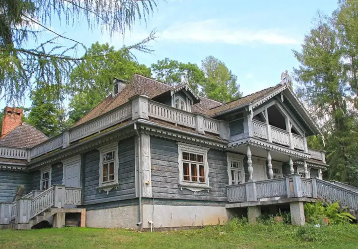 Drewniany dworek w parku pałacowym w Białowieży, z werandami i ozdobnymi detalami, otoczony zielenią.