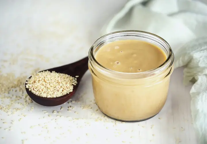 Un vasetto di tahina, crema di sesamo, accanto a un cucchiaio di semi. Scopri cos'è la tahina e come usarla.