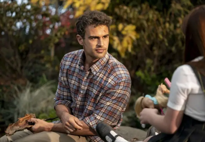 Theo James w kratowanej koszuli, znany z filmów, seriali i programów, siedzi na zewnątrz. Obok niego dziecko bawi się lalką.