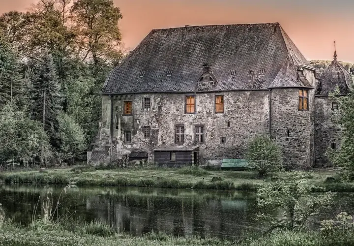 Kamienny polski dwór szlachecki z oknami oświetlonymi ciepłym blaskiem, otoczony zielenią i stawem.