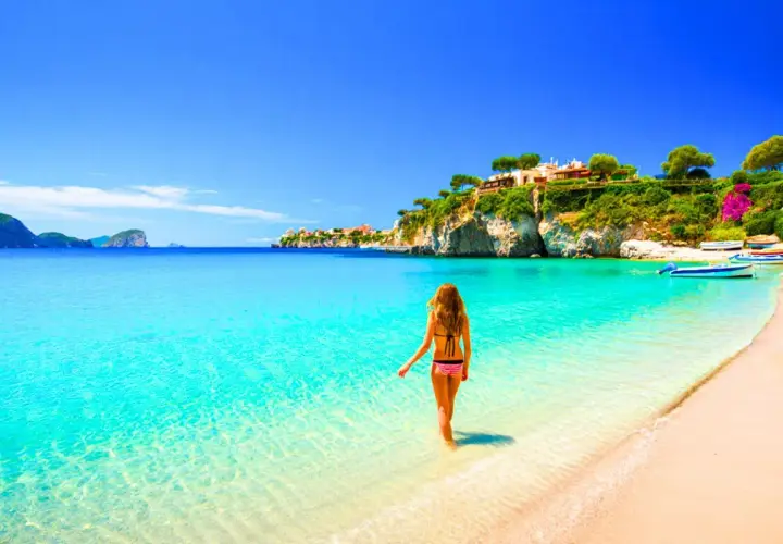 Femme en bikini entrant dans l'eau turquoise d'une plage idyllique, avec des maisons perchées sur une falaise, typique de la côte méditerranée en Italie.