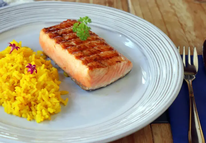 Un delicioso salmón a la plancha con marcas de parrilla, servido con arroz amarillo y decorado con perejil y flores moradas.