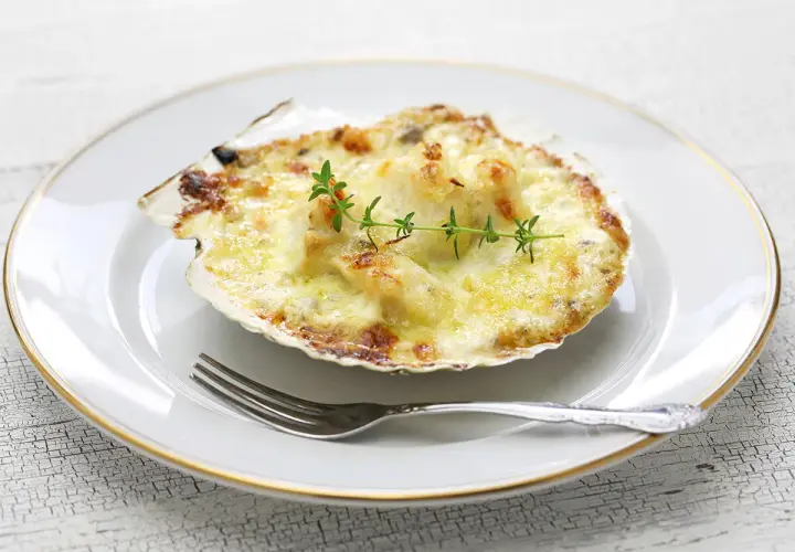 Deliciosas vieiras gratinadas en su concha, cubiertas de queso derretido y doradas, adornadas con una ramita de tomillo.
