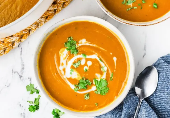 A creamy, vibrant bowl of kabocha squash soup, swirled with coconut cream and garnished with fresh cilantro. A spoon rests on a blue napkin beside it.