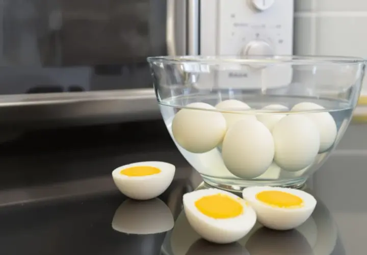 Des œufs durs dans un bol d'eau, prêts à être dégustés. Un œuf est coupé en deux, révélant un jaune d'or parfait. La méthode de cuisson par oeuf dur micro onde est rapide et facile.