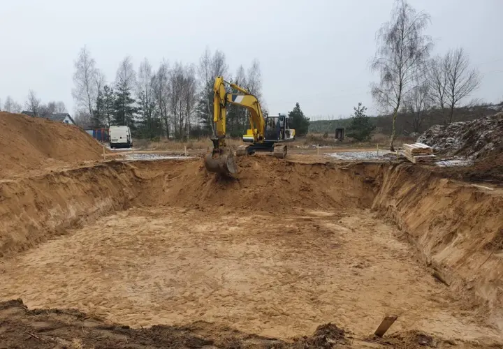 Koparka pracuje przy wykopie pod fundament. Piasek i ziemia tworzą głęboki dół, przygotowany do dalszych prac budowlanych.