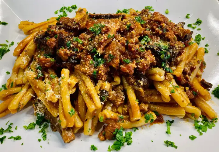 Un piatto di pasta alla norma fumante, con melanzane fritte, pomodoro e ricotta salata grattugiata, guarnito con prezzemolo fresco.