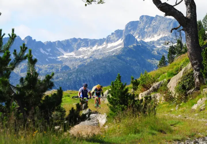 Ciclistas descienden por un sendero en el Pla de Beret, con imponentes montañas nevadas de fondo.