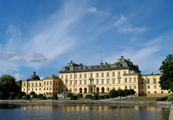 Drottningholm slott, ett gult barockpalats med gröna tak, speglar sig i vattnet under en blå himmel med tunna moln.