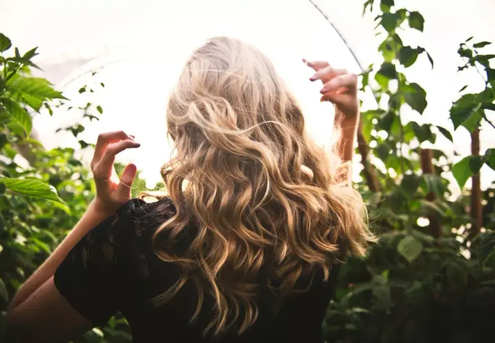 Blond włosy falują na wietrze, jakby przechodziły anwen test na porowatość. Kobieta w czarnej koronkowej bluzce stoi wśród zielonych liści.