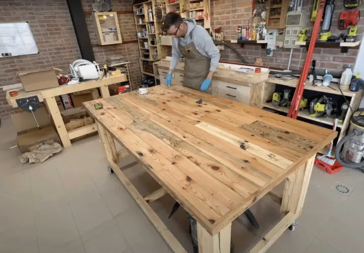 Uomo con guanti blu applica finitura su un robusto banco da lavoro in legno fai da te, circondato da attrezzi e materiali in un laboratorio.