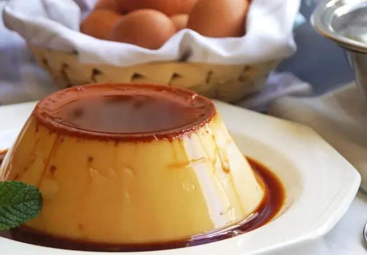 Delicioso flan de huevo casero con caramelo líquido, decorado con menta fresca. En el fondo, una cesta con huevos y un frasco de mermelada.