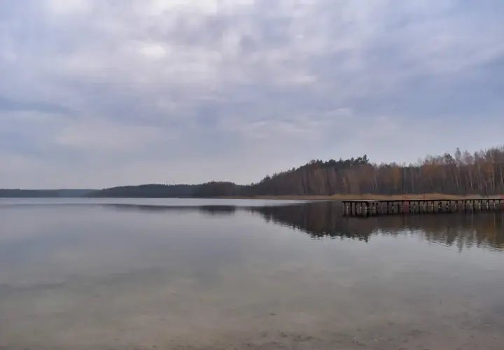 Spokojne jezioro Kamionki odbija pochmurne niebo i lasy. Drewniane molo wchodzi w głąb wody.