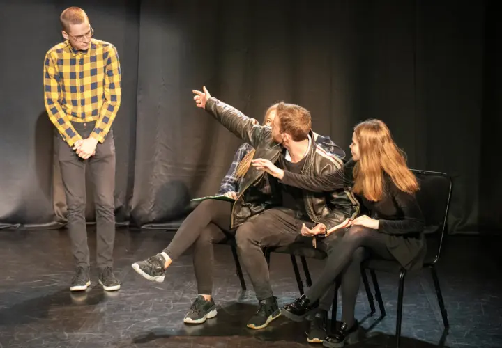 Młodzi aktorzy ćwiczą scenę. Jeden z nich wskazuje coś na widowni, jakby to był teatr na bruku.