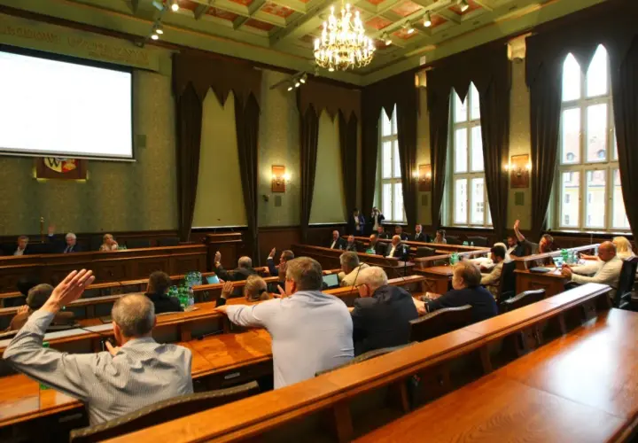 Sala obrad z ludźmi podnoszącymi ręce, widok zza pleców uczestników.