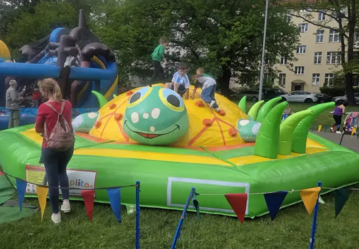Dzieci bawią się na dmuchanym placu zabaw w kształcie gąsienicy. W tle widać inne dmuchańce i budynki.