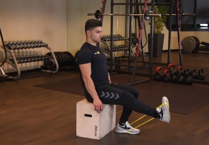 Mężczyzna wykonuje ćwiczenie sissy squat z gumą oporową na nogach, siedząc na skrzyni w siłowni.