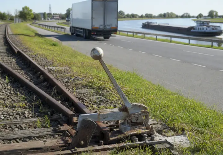 Alte Weiche neben Gleisen. Ein LKW und ein Frachtschiff sind auf der Straße und dem Wasser zu sehen, verschiedene Verkehrsmittel.