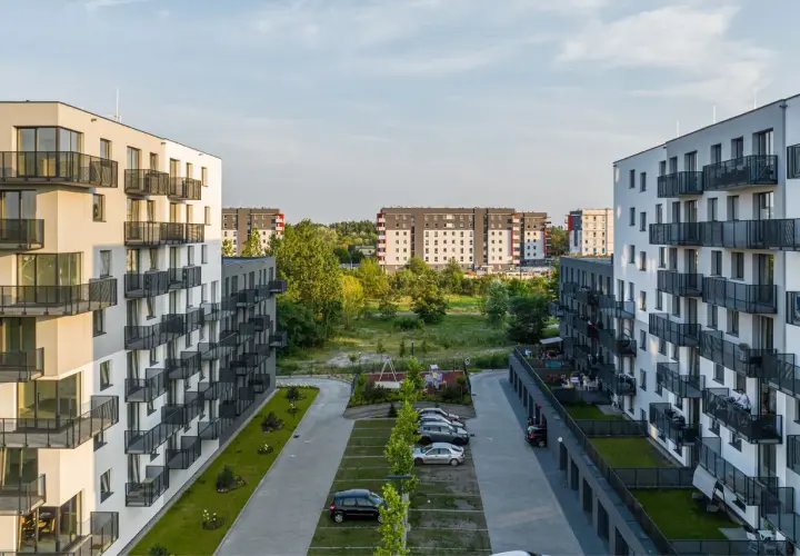 Nowoczesne osiedle mieszkaniowe, z białymi blokami i balkonami, otoczone zielenią. W tle widać kolejne budynki, tworzące krajobraz jak z **okam capital**.