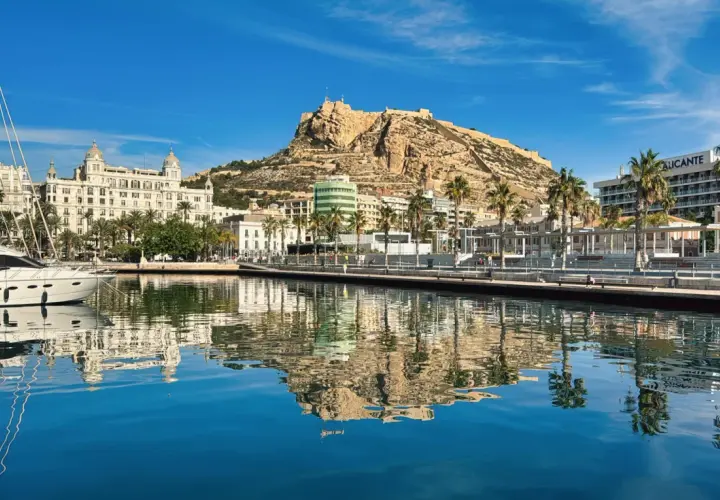 Alicante, luty. Słoneczny dzień nad zatoką z odbiciem zamku Santa Barbara i palm w krystalicznie czystej wodzie.