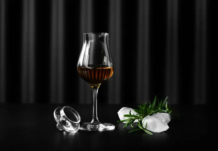 Szklanka Glencairn z bursztynowym trunkiem, obok leży jej zakrętka, lód i rozmaryn. Jak dobrać odpowiednie szkło Glencairn vs tumbler vs copita.