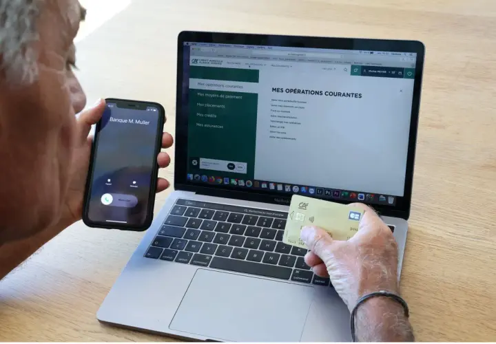 Un homme tient un téléphone et une carte bancaire, devant un ordinateur affichant le site du Crédit Agricole. Il cherche peut-on annuler un paiement par carte bancaire sur internet.